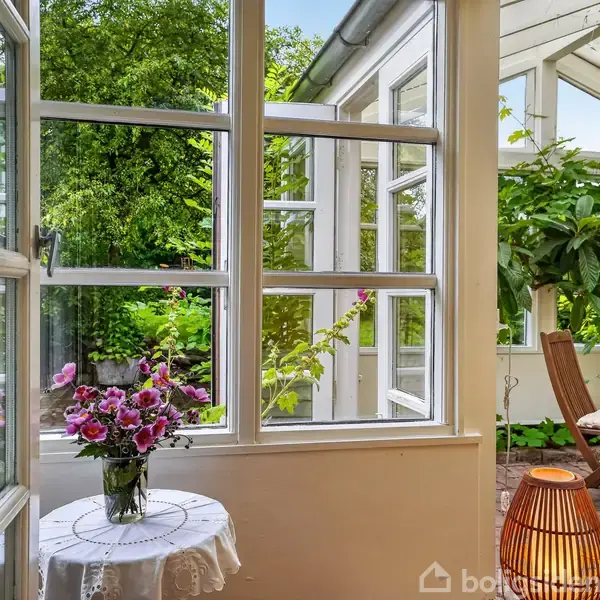 En vase med blomster står på et bord ved et åbent vindue. Haven udenfor bugner af grønne planter, og der er en stol ved et bord i den lyse udestue.