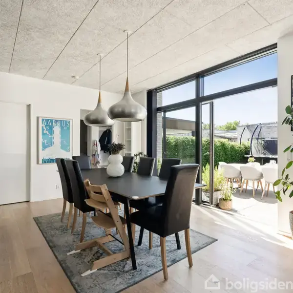 Spisebord med sorte stole står på tæppe i moderne stue, oplyst af store vinduer og dekoreret med planter og kunst. Terrasse med havemøbler ses udenfor.