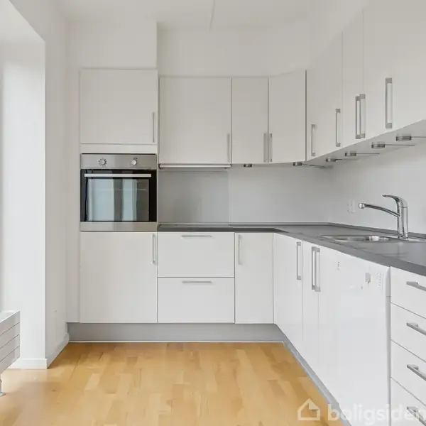 Et moderne køkken med hvide skabe og skuffer rundt om et gråt køkkenbord. Der er en indbygget ovn til venstre og en stor glasdør modsat.