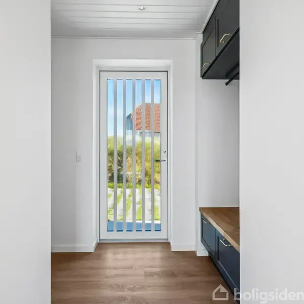 En glasdør med lodrette skodder åbner mod en have, set fra et rum med træparketgulv og mørkeblå skabe. Udenfor ses en græsplæne og et rødt tag.