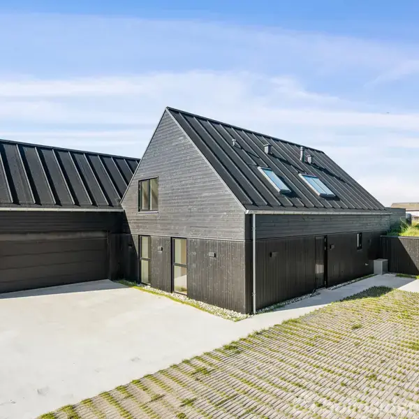 Moderne sortmalet hus med skråtag står på en åben grund med flisebelagt indkørsel; omgivet af græs og en lysende blå himmel.