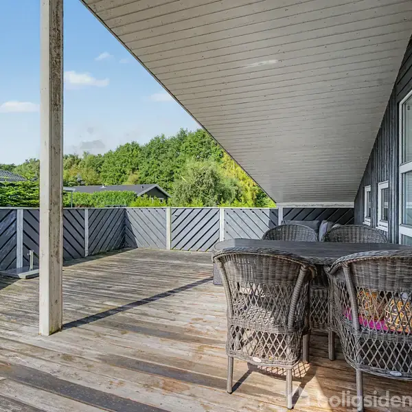 Terrasse med træbord og fletstole under halvtag. Omgivet af træbeklædte afskærmninger, grønne træer i baggrunden. Skyfri himmel skaber en lys og åben atmosfære.