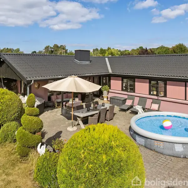 Et hus med sort tag og rosa facade. Terrasse i haven, hvor der står et bord med parasol. Rund pool til højre. Baggrunden er en klar blå himmel med skyer.