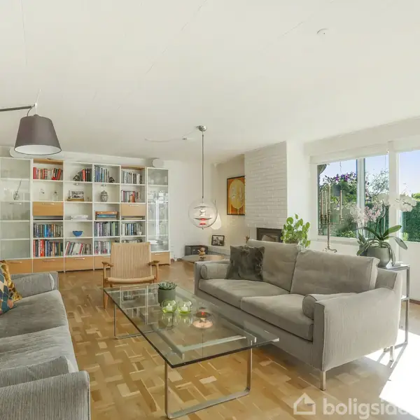 Stue med grå sofaer og glasbord, stående på trægulv. Reol med bøger dominerer bagvæggen. Store vinduer åbner til en græsplæne med hæk udenfor, giver rummelig og lys atmosfære.