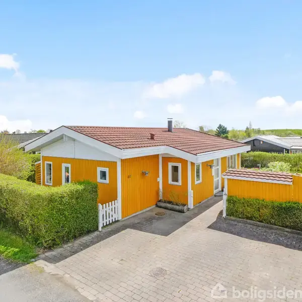 En gul træhusbygning med rødt tag står omgivet af en hæk. Der er en flisebelagt indkørsel foran huset i et fredeligt boligområde med flere huse i baggrunden.