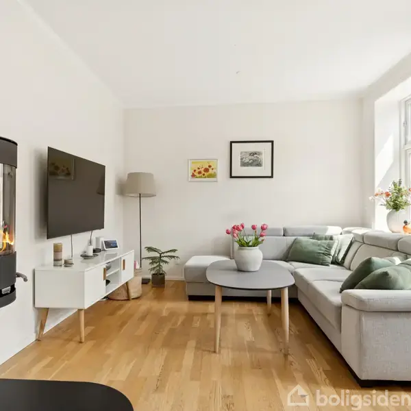 Væghængt pejs med flammer oplyser en stue med en grå hjørnesofa, sofabord og vægkunst. Et fladskærms-tv hænger ved siden af en reol med planter og en lampe.