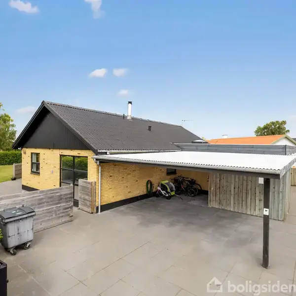 Et gulstenshus med sort tag ses fra gården, hvor en carport med et skur står ved siden af. En græsplæne med gyngestativ er i baggrunden.