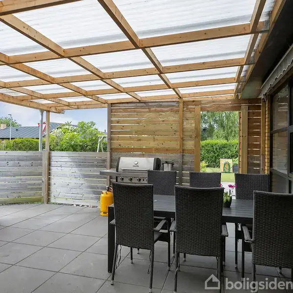 En overdækket terrasse med trærammer og gennemsigtigt tag har et sort spisebord og stole. En grill står ved siden af, omgivet af haveudsigt og planter i baggrunden.