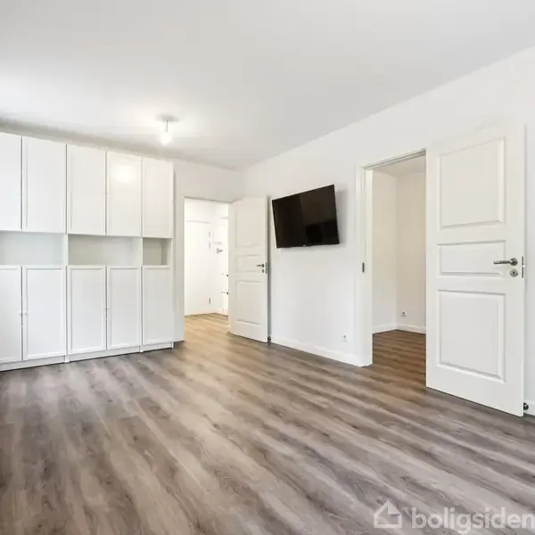 Stue med trælaminatgulv har et væghængt TV, hvide skabe langs bagvæggen. Væggene er hvide, og rummet har to døre og grå gardiner.