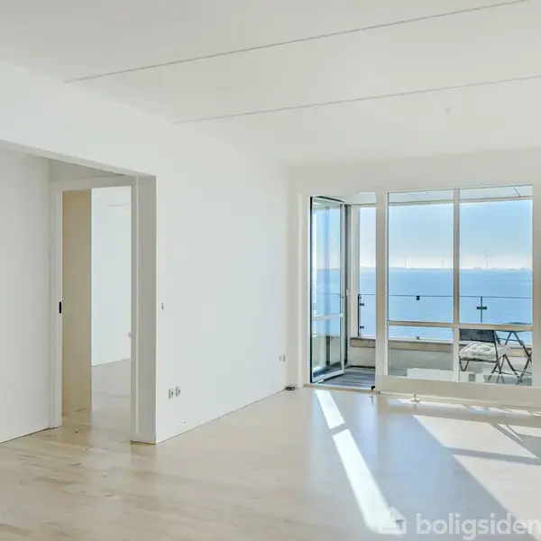 Et lyst rum med trægulv og hvide vægge, åbner sig mod en balkon med udsigt over havet. En enkel stålampe står til højre ved vinduet.