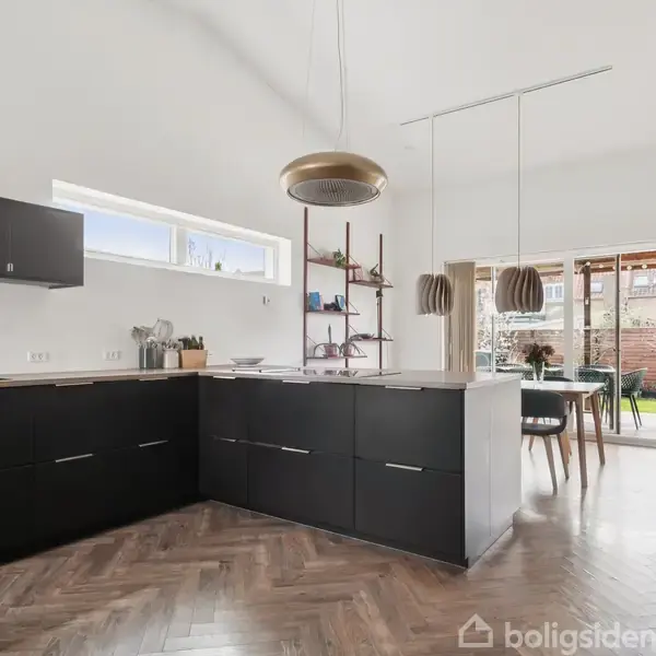 Svart køkken med moderne skabe og skuffer, indbyggede apparater og hvidt bord. Et lille vindue giver naturligt lys. Mørkt trægulv giver en varm atmosfære.