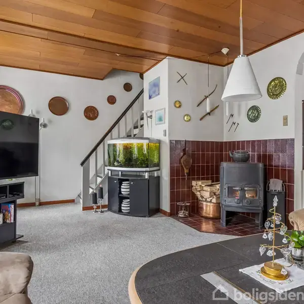 Stue med en stor fladskærms-tv, et akvarium ved trappen og en brændeovn i hjørnet. Træpaneler i loftet og dekorative tallerkner på væggen.