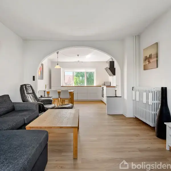 Sofa og lænestol står i en stue med trægulv, sofabord og vægkunst. Rummet åbner op mod et køkken med spisebord, set gennem en buegang.