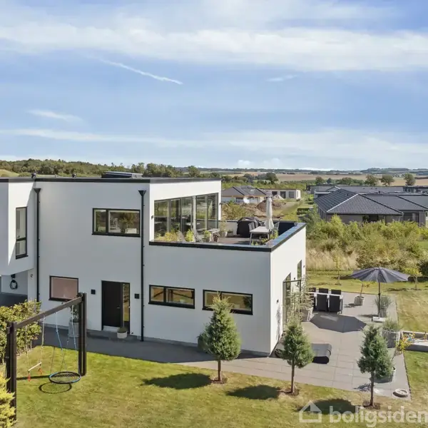 En moderne, hvid villa med fladt tag står på en velplejet græsplæne. Der er en terrasse med havemøbler og parasol, omkranset af buske og træer, i et boligkvarter.