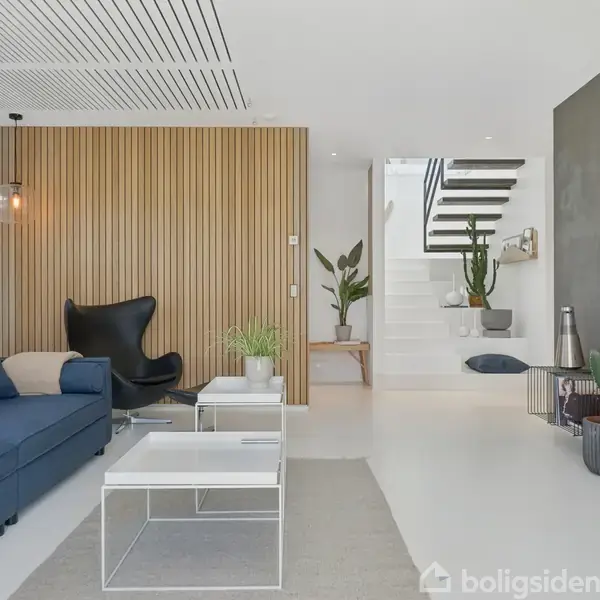 Blå sofa i en moderne stue med træpaneler og sorte stole. Rum med hvide vægge, planter, og en fladskærm. Trappe fører til anden etage.