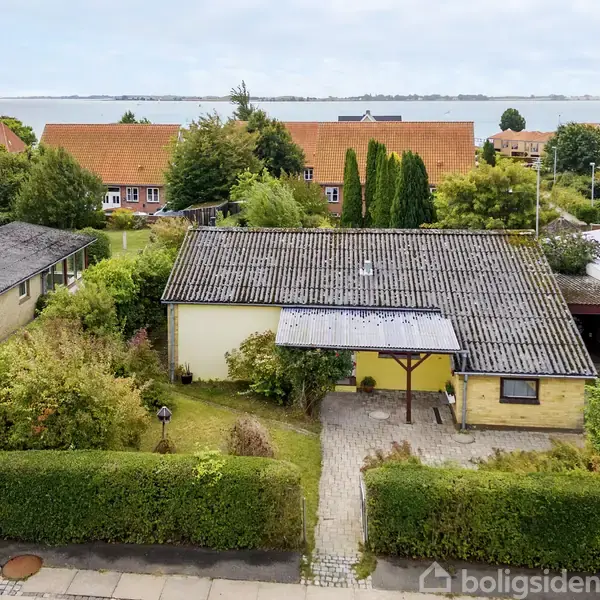 Et gult hus med fladt tag ligger i en villakvarter. Huset er omgivet af en velplejet have med grønne buske. I baggrunden ses flere huse og kystlinjen.