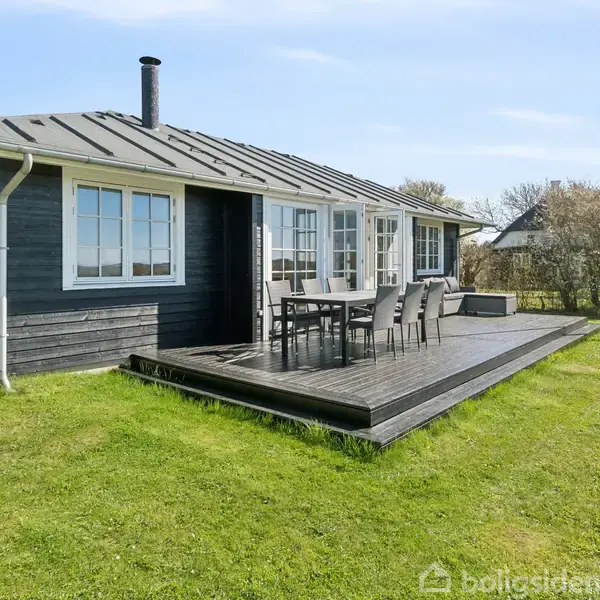 Sort træhus med store vinduer står stille på græsplæne, omkranset af buske. En terrasse med havemøbler ses foran, oplagt til udendørs ophold og hygge.