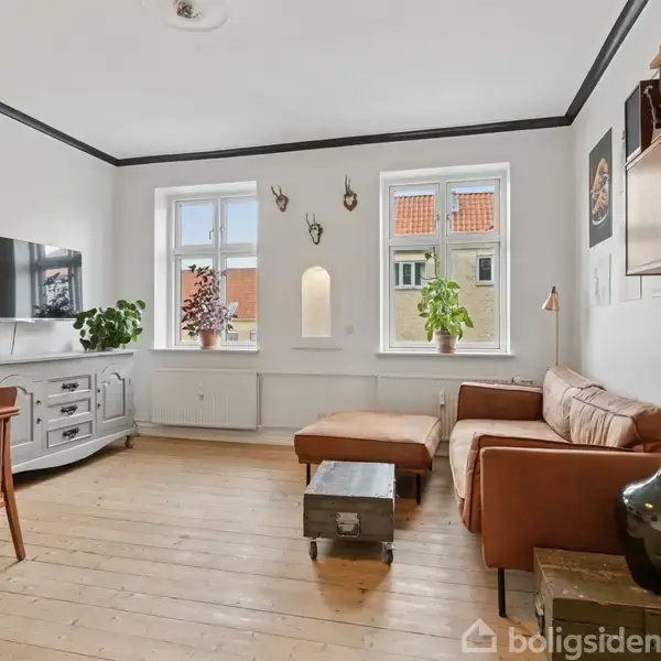 Stue med brunt sofahjørne ved vinduer, fladskærm på væggen, træbord med stole, og flere planter på gulv og vindueskarm. Lyst træparketgulv og hvidmalede vægge.