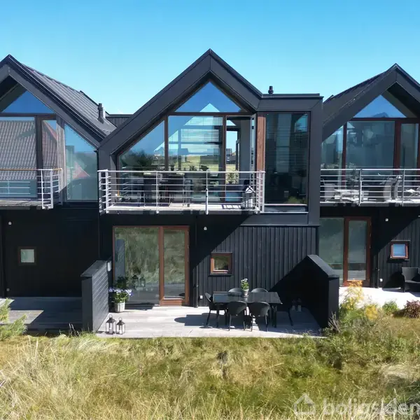 Sort moderne hus med store glaspartier og altaner set udefra. Husets terrasser er omgivet af græs og sandklitter, hvilket skaber en naturskøn beliggenhed. Ingen tekst ud over vandmærket.