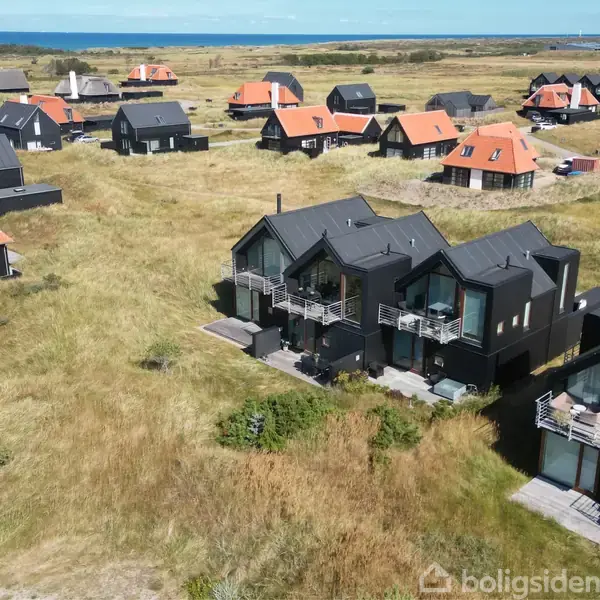 Flere sorte træhuse med skarpe tagkanter står på græsareal ved kysten. Andre huse med orange tage ses omkring dem, og havet er synligt i baggrunden.