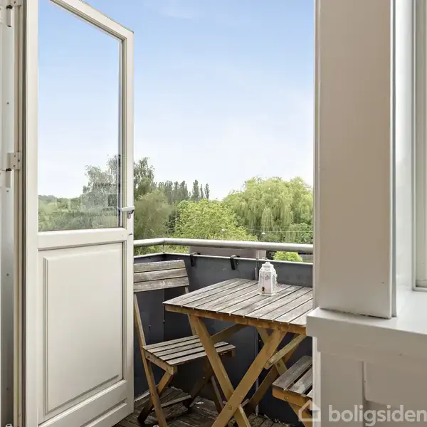 Åben altandør viser en mindre altan med et bord og to stole. Udsigt til grønne træer og blå himmel. Áltanen er omkranset af en lav rækværk.