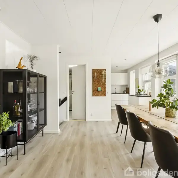 Sort vitrineskab indeholder glas og flasker, står i stue med lyst trægulv. Lang spisebord af træ med sorte stole, planter og pendellampe. Vinduer på højre side.
