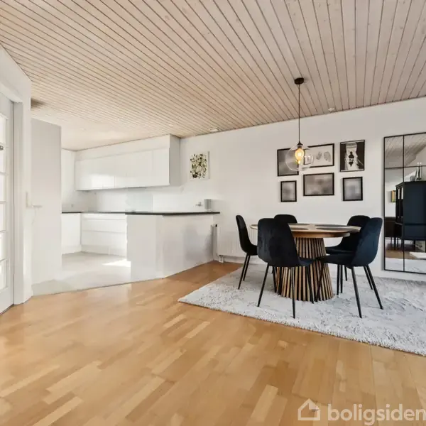 Rundt spisebord med fire sorte stole står på et lyst tæppe i en moderne spisestue. Væggen har flere billeder og et stort spejl. Køkkenområdet er i baggrunden.