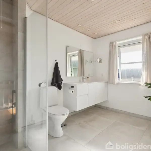 Et badeværelse med et hvidt toilet og en vask isat i et skab. Et spejl hænger over vasken. Rummet har lyse fliser, træloft og et vindue med gardiner. En grøn plante står i hjørnet.