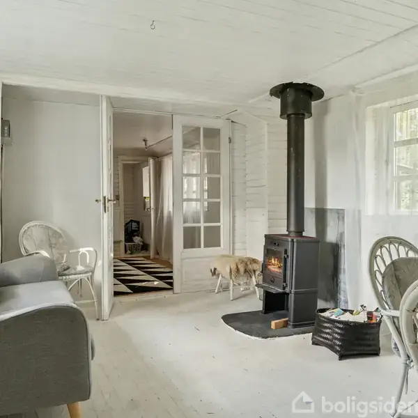 En brændeovn står i et lyst rum med hvide vægge og møbler, herunder en grå sofa og kurvestole. Store vinduer lader naturligt lys strømme ind.