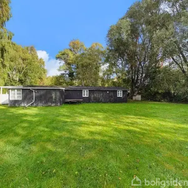 En sort træbygning med hvide vinduer står stille omgivet af en frodig, grøn græsplæne. Store træer omkranser bygningen, og himlen er blå med enkelte skyer.
