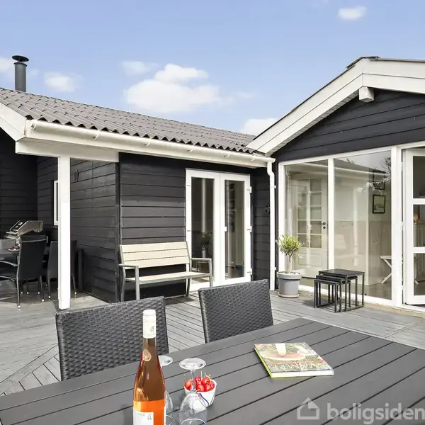 Sortmalet træhus står ud mod en stor træterrasse. På terrassen er der et bord med stole, en flaske vin og et glas. I baggrunden ses blå himmel og omgivende natur.