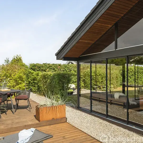 Et moderne hus med store glaspartier reflekterer solen, omgivet af en terrasse med træmøbler og grønne planter i krukker, der giver en charmerende udendørs atmosfære.