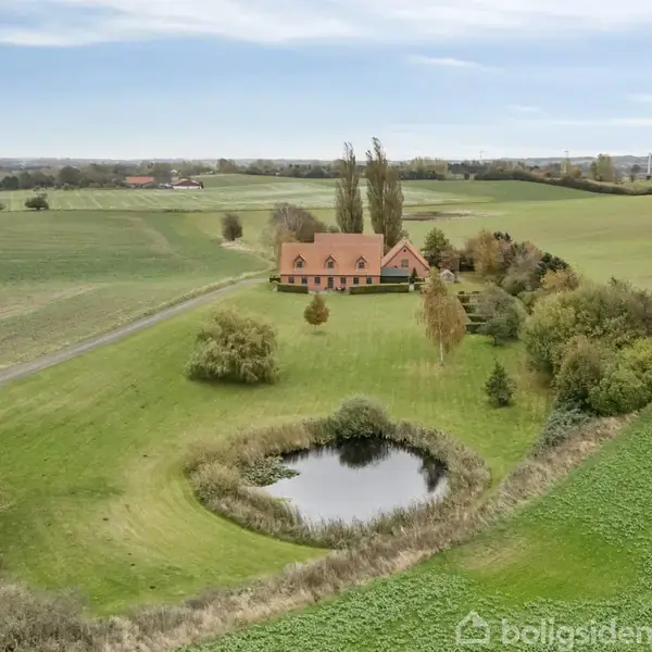En rød villa med skråtag står stille i en frodig, landlig setting. Foran huset er der en rund dam omgivet af grønne marker og spredte træer.