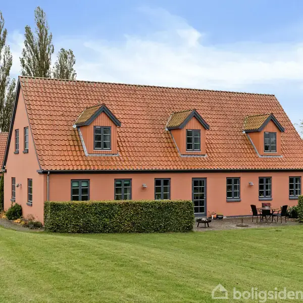 Et stort, pink hus med rødt tegltag har flere kviste og vinduer. Det står på en velplejet græsplæne omkranset af hække. To høje træer ses i baggrunden.