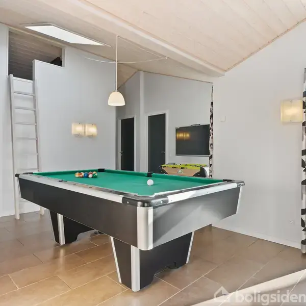 Et billardbord står klar til spil i et moderne, lyst rum med lyse vægge og store vinduer. En stige fører op til en hems. Der er ingen tekst.