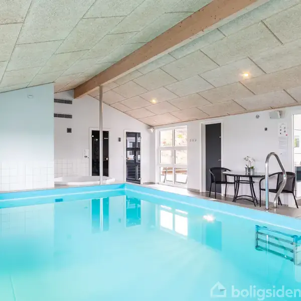 Indendørs swimmingpool står stille i et lysegråt rum med et lavt loft. Der er en lille bord- og stolarrangement ved siden af poolen og flere døre fører ud til andre rum.