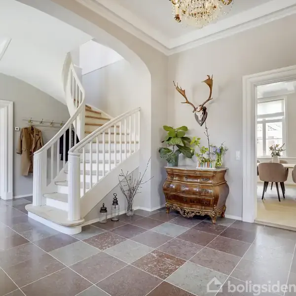 Spiraltrappe fører opad i en elegant entré med lyse fliser, en antik kommode med planter og hjortetrofæ. Til højre ses et spiseområde med lyse møbler og stor lampe.