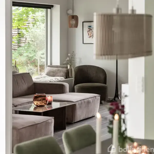 En moderne stue med en grå sofa og glasbord, hvorpå der står en kobberfarvet skål og stearinlys. Store vinduer giver udsigt til en grøn have.