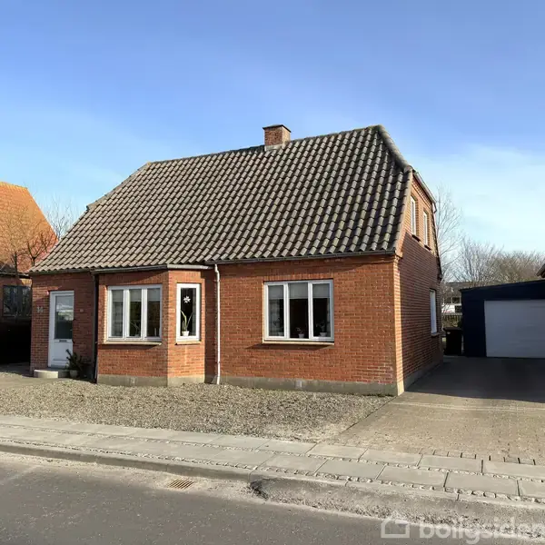 Rødstenshus med mørkt tegltag står på en gade, omgivet af andre huse. En indkørsel fører til en garage i højre side, og der er ingen tekst.