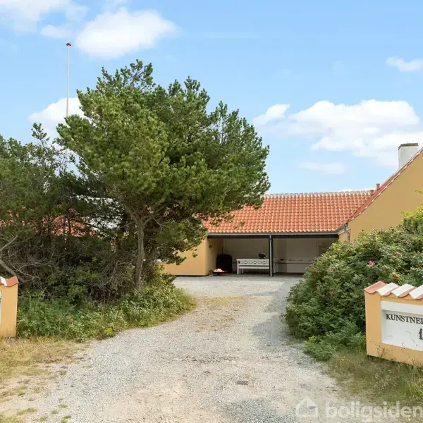 Bygning med rødt tegltag står stille, omgivet af træer og buske. Grusvej fører op til huset. To skilte ved indgangen har teksten "KUNSTNERGAARDEN 10".