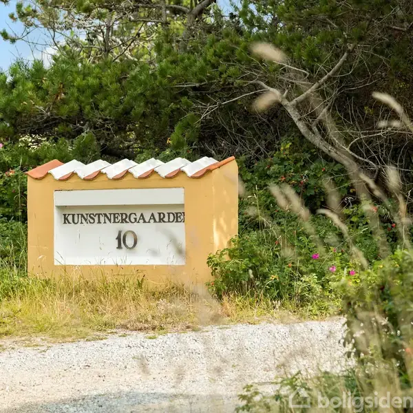En skilt står ved en grusvej og viser teksten "KUNSTNERGAARDEN 10" omgivet af frodig vegetation og træer under en klar blå himmel.
