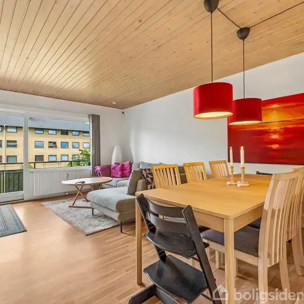 Stue med spisebord og seks stole, hvorpå der står lysestager. En sofa er placeret ved et vindue med udsigt til en bygning. Loftslamper hænger over bordet.