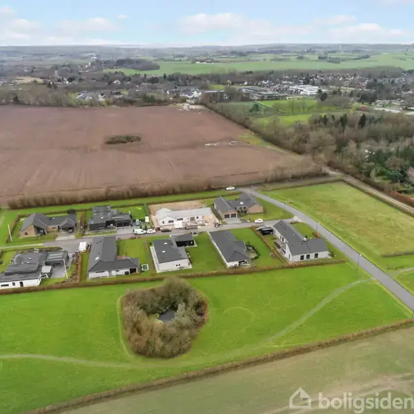 Boligområde med moderne huse placeret på række; omkranset af grønt græsareal og marker; beliggende nær en vej. I baggrunden er der skove og en by med spredte bygninger.