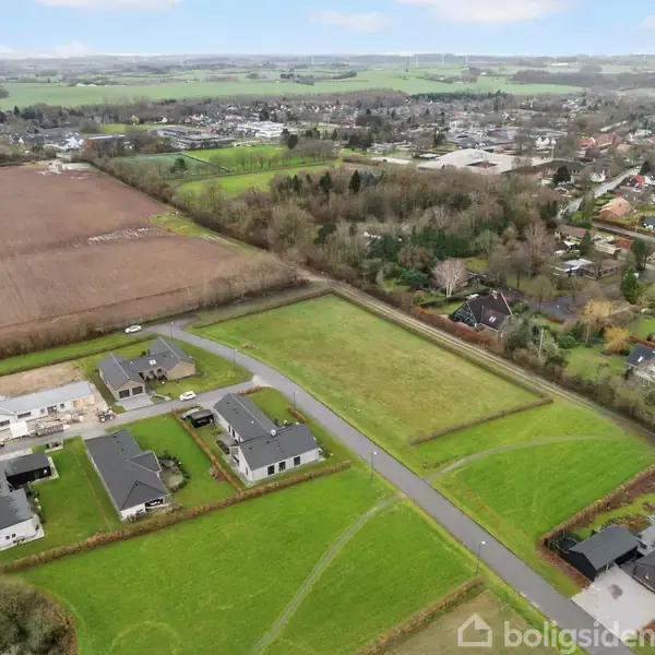 Nogle huse er placeret langs en vej under opførelse, omgivet af grønne marker og en landsby i baggrunden, hvilket skaber en landlig og fredfyldt atmosfære.