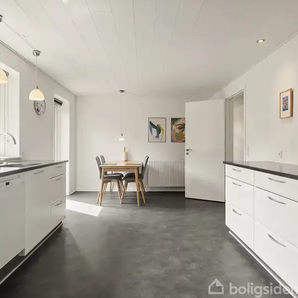 Et moderne køkken med hvide skabe og sort bordplade. Et spisebord med stole står i baggrunden. Billeder hænger på væggen, og sollys strømmer ind fra vinduet.