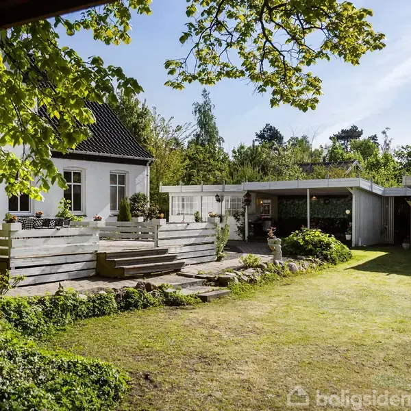 Et hvidt hus med en terrasse og trapper. Det ligger i en frodig have med græsplæne og træer omkransende. Terrasseområder er dekoreret med planter og blomster.