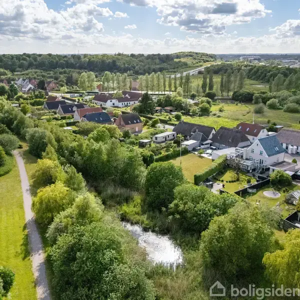 En række parcelhuse ligger stille blandt frodigt grønt landskab og spredte træer, hvor en lille sø kan ses i midten, under en klar himmel med spredte skyer.