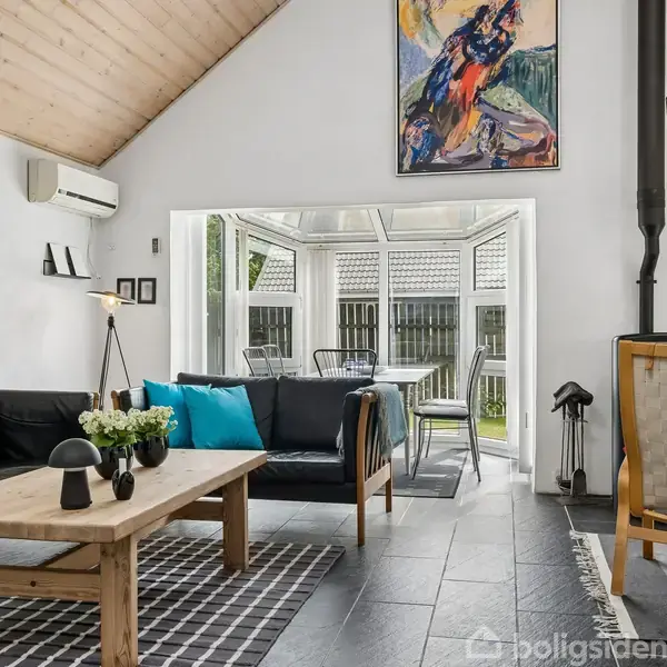 Stue med sort lædersofa og træbord på gråt gulvtæppe. En farverig kunstmaleri på væggen. Vinduer åbner til en terrasse med borde og stole. Væggene er hvide.