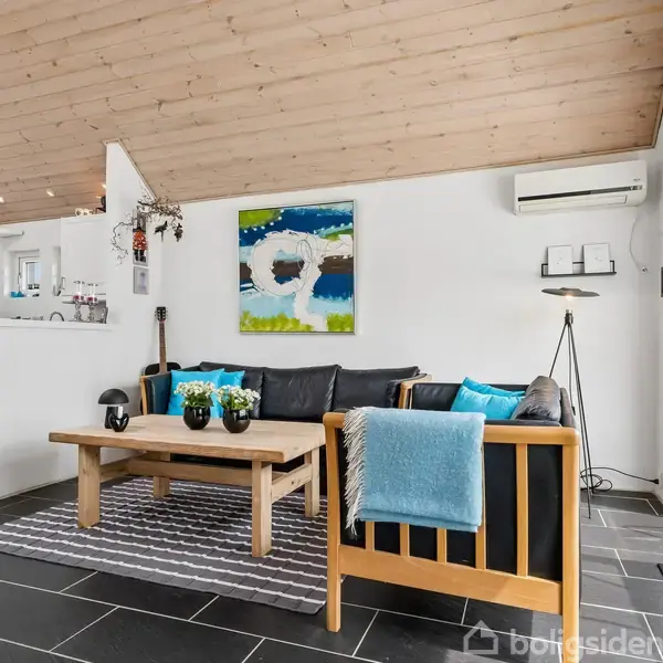 Lys stue med træloft, sort flisegulv og en sort sofa med farverige puder. Et træbord står foran sofaen, og en abstrakt maleri hænger på væggen. Køkken ses i baggrunden.