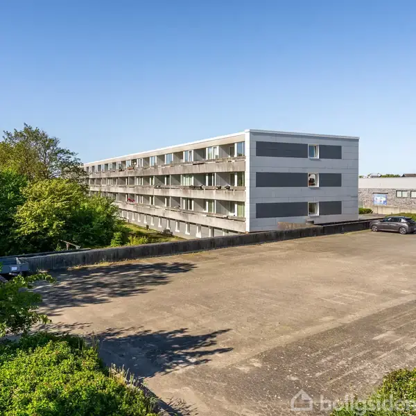 En lang, grå lejlighedsbygning med balkoner strækker sig langs venstre side, mens et stort, åbent parkeringsområde ligger foran, omgivet af grønne træer og blå himmel.